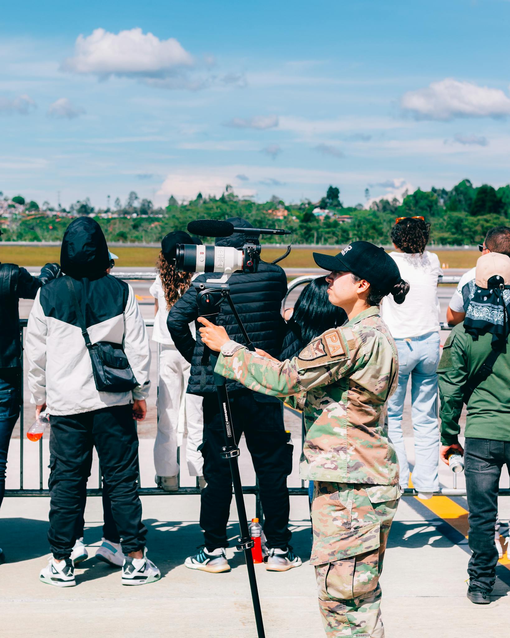 A soldier operates a camera on a monopod while a crowd observes an outdoor scene.